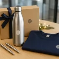 Regali aziendali di Natale: borraccia termica, penne e maglietta personalizzate su un tavolo con decorazioni natalizie